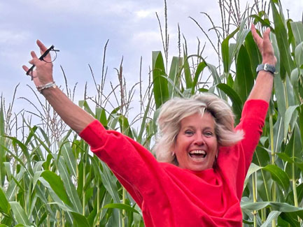 Vrolijke vrouw in rood trui springt met armen omhoog tussen hoge maïsplanten op zonnige dag, glimlachend gezicht