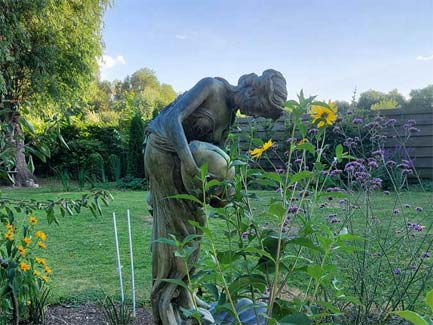 Standbeeld van een vrouwenfiguur met vaas in een weelderige tuin bij zonsondergang, bloemen en groen op de voorgrond