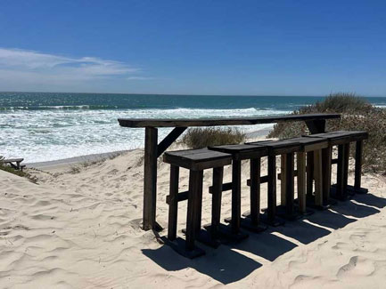 Houten bar en krukken op zonnig strand bij draaipoort lodge met zandduinen en blauwe zee op de achtergrond