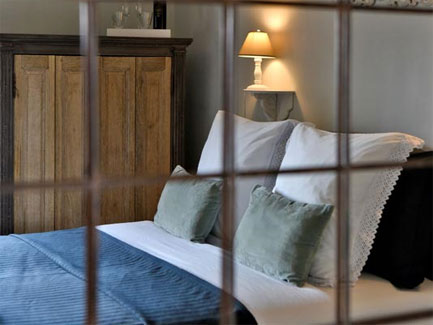 Sfeervolle slaapkamer met wit linnen, lichtblauwe sprei, groene kussens en warme lamp naast houten kast in landelijke stijl