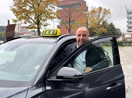 Taxichauffeur stapt uit zwarte taxi bij kade met gele TAXI-daklamp in stedelijke omgeving, herfstbomen op de achtergrond